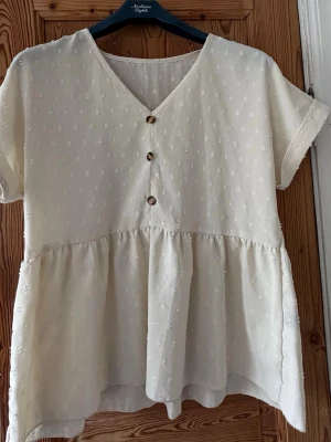 Ljusbeige blus med knappar och struktur - Supersöt ljusbeige blus med v-ringning, korta ärmar och tre dekorativa knappar framtill. Blusen har ett subtilt broderat mönster och volang nedtill för en luftig känsla. Perfekt till jeans eller kjol för en chill look.