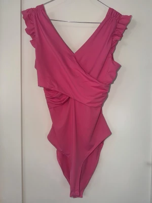 Rosa body/baddräkt med volangärmar - Snygg rosa body/baddräkt med omlott-design och djup V-ringning både fram och bak. Volangdetaljer vid axlarna ger en extra feminin touch. Tillverkad i stretchigt material som sitter bekvämt och smickrande. Perfekt för strand eller poolhäng. Eller som en topp. 