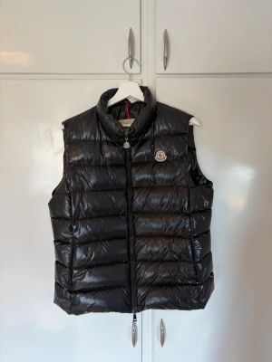 Moncler Ghany gilet  - Snygg svart dunväst från Moncler med glansig finish och klassisk logotyp på bröstet. Västen har hög krage, två fickor med dragkedja och quiltad design. Perfekt för lager-på-lager och streetstyle.