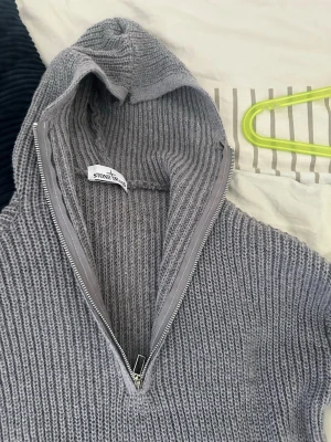 Grå stickad hoodie från Stone Island - Säljer en grå stickad hoodie från Stone Island med dragkedja framtill. Tröjan har en ribbad struktur, huva och långa ärmar. Perfekt för dig som gillar streetwear och vill ha något både snyggt och bekvämt.