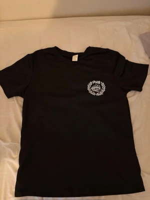 Svart t-shirt med PHS-tryck - Svart t-shirt med vitt PHS-tryck och kransdetalj på bröstet. Klassisk passform och rund halsringning, perfekt för en avslappnad stil.