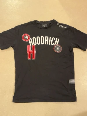 Svart Hoodrich t-shirt med tryck - Svart t-shirt från Hoodrich med stora vita bokstäver och röda detaljer framtill. Flera broderade patches och tryck, bland annat ett stort H och logotyp på bröstet. Rund halsringning och korta ärmar. Skön och stilren streetwear-look.