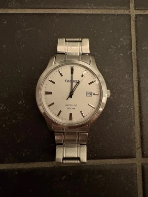 Seiko klocka - Stilren Seiko klocka i silver med länkarmband i metall och vit urtavla. Klockan har datumvisning och lysande indexmarkeringar som syns i mörker. Saphirglas och vattentät upp till 100 meter. Perfekt accessoar för dig som gillar klassisk design.