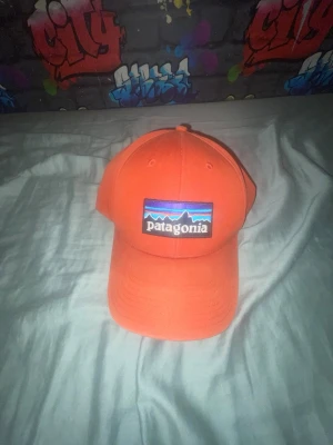 Orange keps från Patagonia - Snygg orange keps från Patagonia med klassisk böjd skärm och justerbar snapback. Framtill sitter en broderad logotyp med bergsmotiv i blått, lila och svart. Kepsen är tillverkad i bomullstyg och har ventilerande hål upptill.