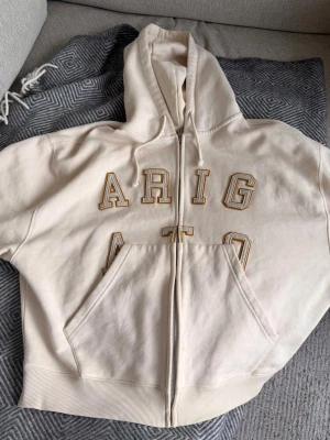 Beige zip hoodie från Arigato - Säljer en beige kofta med dragkedja från Axel Arigato. Koftan har stora broderade bokstäver i gult och vitt på bröstet, huva med dragsko och en stor magficka. Tillverkad i mjuk bomull för skön känsla och avslappnad stil. Nypris: 2000+