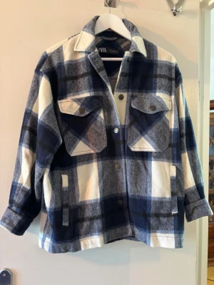Blåvit rutig overshirt från Zara - Overshirt från Zara i blått, vitt och grått rutmönster. Modellen har två stora bröstfickor med lock och knappar, samt sidofickor. Overshirten är i mjukt, tjockt tyg och har avslappnad passform med klassisk krage och tryckknappar framtill. En svart fläck på armen, syns på ena bilden.