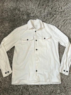Vit overshirt jack and Jones  - Stilren vit overshirt med svarta knappar och två bröstfickor med lock. Långärmad modell med klassisk krage och knappstängning vid ärmslut. Perfekt att bära över en t-shirt för en avslappnad look.