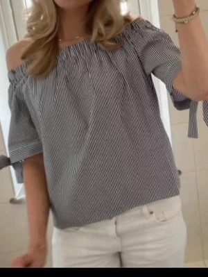Randig offshoulder topp med knyt - Superfin offshoulder topp från Anna field med smala svarta och vita ränder. Toppen har korta ärmar med knyt i ärmsluten och en avslappnad passform. Perfekt för en trendig och somrig look.
