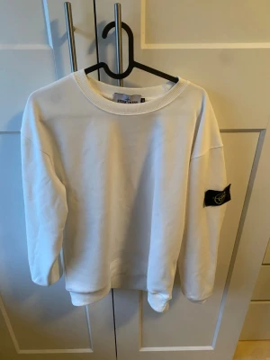 Vit sweatshirt från Stone Island - Säljer en clean vit sweatshirt från Stone Island med klassisk logga på ärmen. Tröjan har rund halsringning, ribbade muddar och är gjord i mjukt material. Perfekt för en stilren och avslappnad look.