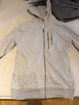 Ljusgrå hoodie från Sail Racing - Ljusgrå hoodie med dragkedja från Sail Racing. Hoodien har en bröstficka med dragkedja och broderad logga på vänster sida. Mjuk bomullsblandning, ribbade muddar och stor huva. Perfekt för en avslappnad och sportig stil.
