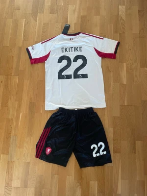 Liverpool matchställ Ekitike nr 22 - Säljer ett Liverpool matchställ med Ekitike och nummer 22. Tröjan är vit med röda och svarta detaljer, klubbmärke och sponsortryck framtill. Shortsen är svarta med röda ränder och nummer 22. Perfekt för fotbollsträning eller match.                 Passar kille ca 187cm.
