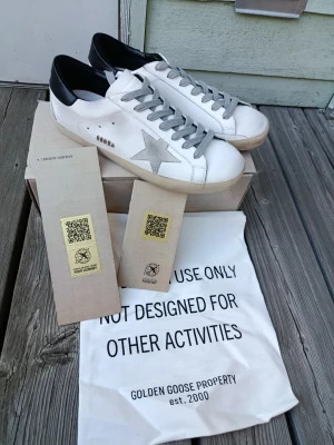 Golden Goose vita sneakers med stjärna - Golden Goose sneakers i vitt läder med grå mockadetaljer och silverfärgad stjärna på sidan. Skorna har grå skosnören, svart hälkappa och klassisk låg siluett. Snygg och ikonisk design med märkets logga på plösen och sidan.