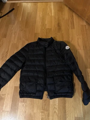Svart dunjacka från Moncler - Snygg svart dunjacka från Moncler med quiltat mönster, två stora fickor framtill och dragkedja. Klassisk Moncler-logga på ärmen och elastiska muddar. Perfekt för kalla vinterdagar och riktigt stilren look.