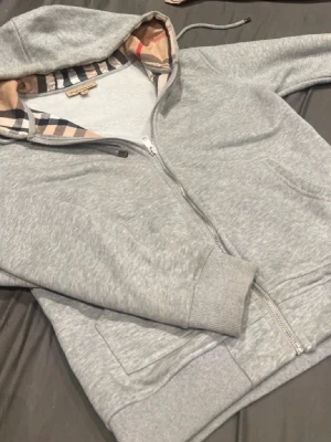 Grå zip-hoodie från Burberry - Snygg grå hoodie med dragkedja från Burberry. Klassisk huva med det ikoniska rutmönstret på insidan. Mjukt material och ribbade muddar vid ärmslut och nederkant. Perfekt för en chill och stilren look.