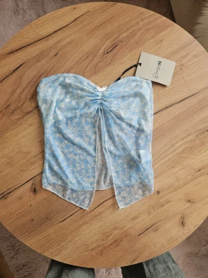 Blå bandeau tubtopp från Design by si - Söt bandeau tubtopp från Design by sii i ljusblått med vitt blommigt mönster. Toppen är ärmlös och har en snygg slits framtill samt rynkdetalj vid bysten. Tunt och lätt material, perfekt för varma dagar.