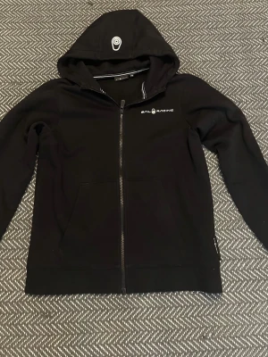 Svart hoodie från Sail Racing - Svart hoodie från Sail Racing med dragkedja framtill och vit logga på bröstet. Huva med tryckt märke och vita kontrastsömmar på insidan. Praktisk känguruficka och mjukt material som känns skönt mot huden. Perfekt för en avslappnad och ren stil