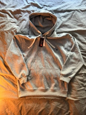 Grå hoodie från Ralph Lauren - Snygg grå hoodie från Ralph Lauren med klassisk huva och vita dragsnören. Har en liten broderad logga på bröstet och en stor magficka framtill. Perfekt för en chill och stilren look. Tillverkad i mjukt material för extra komfort. Dem är lite korta i ärmarna