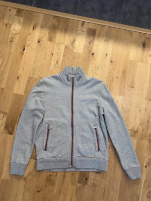 Grå cardigan från Moncler - Snygg grå ziptröja från Moncler med hög krage och dragkedja framtill. Tröjan har två fickor med dragkedja och randiga detaljer i rött, vitt och blått. Liten Moncler-logga på bröstet. Perfekt för en avslappnad och stilren look.