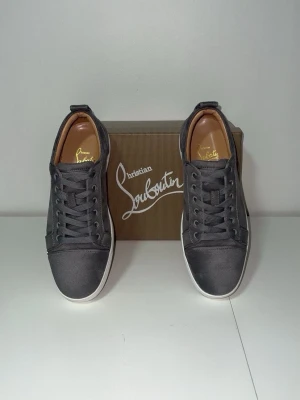 Gråa Christian Louboutin sneakers - Snygga gråa sneakers från Christian Louboutin med klassisk låg siluett, vita sulor och den ikoniska röda undersidan. Skorna har ovandel i mocka och detaljer i läder, samt matchande gråa skosnören. Perfekta för dig som vill sticka ut med stil.