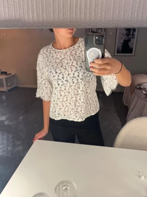 Vit spetsblus med vida ärmar - Säljer en vit blus i spets med blommigt mönster och vida, trekvartsärmar. Toppen har rund halsringning och avslappnad passform. Perfekt att styla med jeans eller kjol för en trendig look.