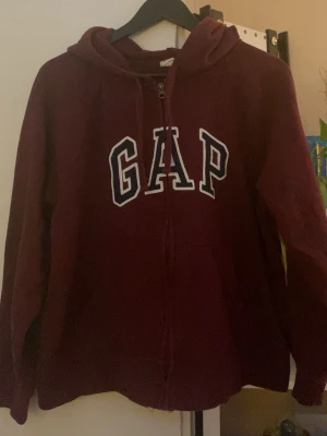 GAP zip up hoodie - Säljer en vinröd hoodie från GAP med dragkedja och stor vit logga framtill. Hoodien har huva med dragsko och är tillverkad i mjukt material, perfekt för en avslappnad stil.