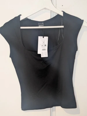 Svart topp från Gina Tricot - En enkel och stilren svart topp från Gina Tricot med rundad halsringning och korta ärmar. Toppen har en tight passform och är tillverkad i ett mjukt stretchigt material som sitter snyggt på kroppen. Perfekt att matcha med jeans eller kjol.