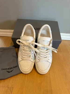 Vita sneakers från Zadig & Voltaire - Snygga vita sneakers från Zadig & Voltaire i läder med perforerade detaljer och blixtdesign på sidan. Klassisk låg modell med vita skosnören och diskret branding på plösen. Använda endast ett fåtal gånger. Nypris 3450kr.