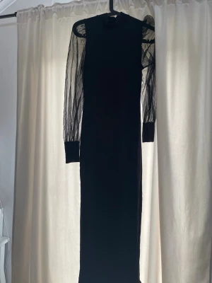 Svart långklänning med meshärmar - Snygg svart långklänning med transparenta meshärmar och höga muddar. Klänningen har en rak passform och högt halsringning, vilket ger en stilren och modern look. Perfekt för dig som vill sticka ut med en elegant och trendig stil.