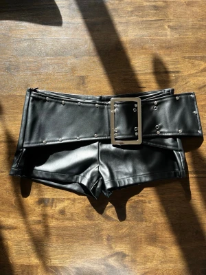 Svarta shorts i skinnimitation med bälte - Säljer ett par svarta shorts i skinnimitation med hög midja och ett brett bälte med stora nitar och metallspänne.