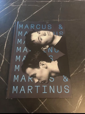 Marcus & Martinus bok - En bok med hård pärm och svart omslag med blå text och porträtt av Marcus & Martinus på framsidan. Boken är på svenska och innehåller berättelser och bilder om popduon. Perfekt för fans som vill veta mer om deras liv och karriär.