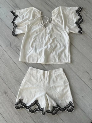 Vitt set med brodyr från Vero Moda - Säljer ett vitt tvådelat set från Vero Moda med topp och shorts. Setet har svarta broderade detaljer längs kanterna och vida ärmar på toppen. Materialet är tunt och luftigt, perfekt för varma dagar. Toppen har knytning vid halsen och båda delarna har en avslappnad passform.