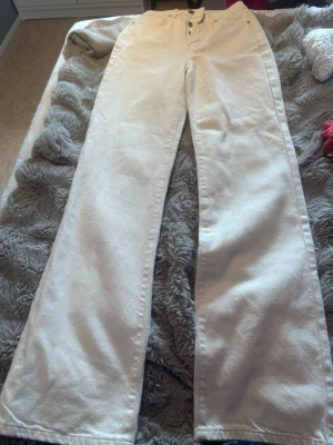 Vida vita jeans med hög midja - Säljer ett par vita jeans med hög midja och raka, vida ben. Klassisk femficksmodell med knappgylf och silverfärgade nitar. Perfekta för en clean och trendig look. Materialet är kraftigt denim i bomull.