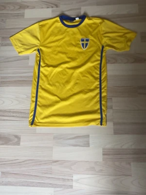 Gul Sverige fotbollströja - Säljer en gul fotbollströja med blå detaljer och Sveriges landslagssköld på bröstet. På ryggen står det 'SWEDEN' i stora bokstäver. Tröjan har rund hals och är tillverkad i ett lätt och snabbtorkande material, perfekt för träning eller match.