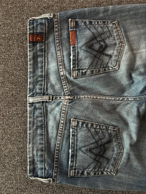 Bootcut jeans från 7 For All Mankind - Snygga ljusblå bootcut jeans från 7 For All Mankind med dekorativa sömmar och små nitar på bakfickorna. Klassisk femficksmodell i mjuk denim med slitna detaljer. Perfekta för en avslappnad och trendig look. Fickorna har paljett liknande detaljer. Strl W26. Super snygga, går ej att köpa nya. 