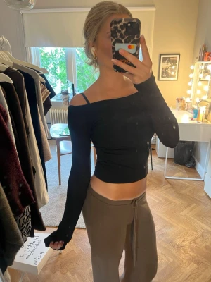 Svart offshoulder croppad topp - Trendig svart offshoulder topp med lång ärm och croppad passform. Toppen har en asymmetrisk axel med ena sidan axelbandslös och den andra med smalt axelband. Perfekt för dig som gillar en edgy och modern stil. Har ett hål på ena sidan av midjan men inget som märks speciellt mycket. Går lätt att laga om man vill det 