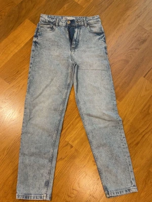 Ljusblå mom jeans från NA-KD - Säljer ett par ljusblå mom jeans från NA-KD med hög midja och raka ben. Jeansen har klassisk femficksdesign, bälteshällor och knappgylf. Materialet är denim i 100% bomull med en snygg tvättad look.