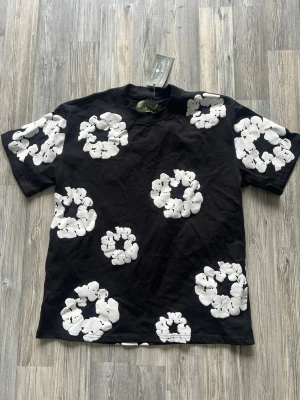 Svart t-shirt denim tears  - Snygg svart t-shirt med stora vita blommiga tryck över hela tröjan. Klassisk rund hals och korta ärmar. Perfekt för dig som vill sticka ut med ett coolt mönster. Tillverkad i mjuk bomull för skön känsla.