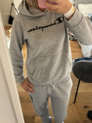 Grått mjukisbyxor och hoodie sett från Champion - Säljer gråt mjukisbyxor och hoodie sett från Champion med logga på benet och bröstet. Byxorna har resår i midjan med dragsko och är gjorda i mjukt sweatshirtmaterial. Perfekt sett för chill dagar eller när du vill vara bekväm. 
