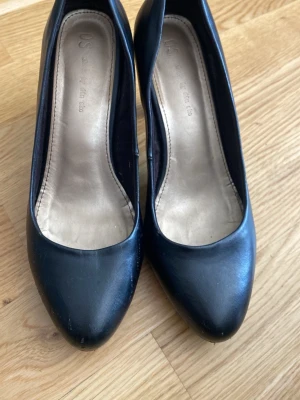 Svarta pumps från Din Sko - Klassiska svarta pumps från Din Sko med rundad tå och låg klack. Skorna har en stilren design och är tillverkade i ett slätt material med beige innersula. Perfekta för dig som vill ha en enkel och tidlös look.