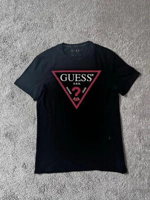 Svart t-shirt från Guess med logga - Snygg svart t-shirt från Guess med den klassiska triangelloggan i rött och vitt på bröstet. Modellen har rund hals och korta ärmar. Perfekt för dig som gillar stilrena och ikoniska plagg med streetkänsla.