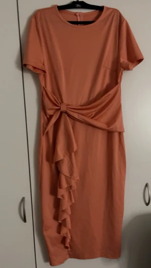 Persikofärgad klänning med volang - Säljer en persikofärgad klänning med knyt i midjan och volangdetalj framtill. Klänningen har rund halsringning och korta ärmar. Perfekt för dig som gillar en feminin och trendig look.