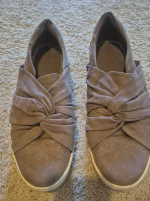 Gammelrosa slip-on sneakers med knutdetalj - Säljer ett par gammelrosa slip-on sneakers med unik knutdetalj på ovandelen. Skorna har rund tå, vit sula och är tillverkade i ett mjukt tygmaterial. Perfekta för dig som vill ha en stilren men ändå trendig look. Använda fåtalet gånger.