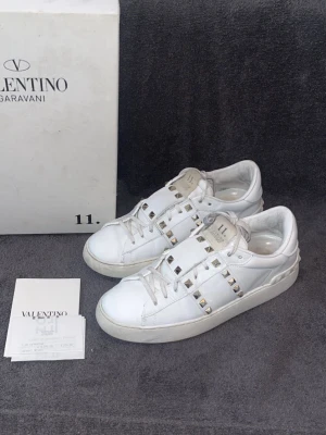 Vita sneakers med nitar från Valentino - Säljer ett par vita sneakers från Valentino Garavani med coola silverfärgade nitar och snörning. Skorna är i skinn och har en stilren, chunky sula. Perfekta för dig som vill ha en edgy touch på din outfit.