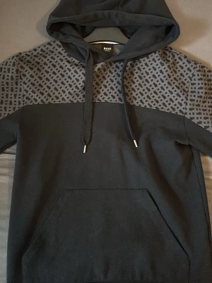 Svart hoodie från BOSS med mönster - Snygg svart hoodie från BOSS med grått geometriskt mönster över axlar och ärmar. Klassisk huva med dragsko och stor magficka framtill. Tillverkad i mjukt material som känns skönt mot huden. Perfekt för en avslappnad och trendig look.