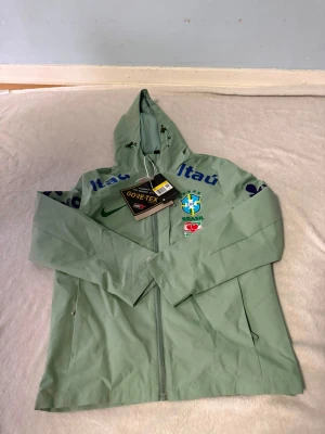 Nike Brasil vindjacka  grön - Ljusgrön vindjacka från Nike med Brasil-logga och sponsortryck. Jackan har huva med dragsko, hel dragkedja och är tillverkad i GORE-TEX-material som står emot regn och vind. Perfekt för dig som gillar sportig stil och vill visa stöd för Brasilien.