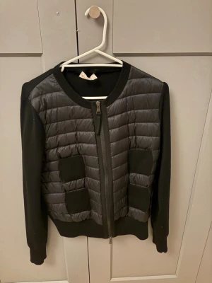Moncler cardigan dam - Snygg svart cardiganjacka från Moncler med quiltad framsida och stickade ärmar. Jackan har rund halsringning, dragkedja framtill och två fickor med ribbade detaljer. Klassisk Moncler-logga på ärmen. Har typ aldrig använt och köpte för 10,245 tusen kronor 