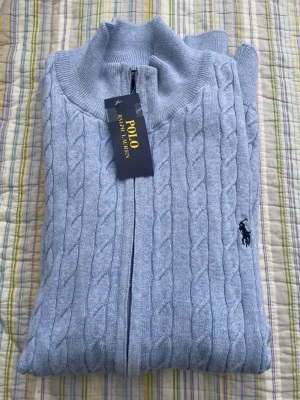 Ljusblå kabelstickad ziptröja Polo - Ljusblå kabelstickad ziptröja från Polo Ralph Lauren med hög krage och dragkedja framtill. Tröjan har det klassiska broderade logotypen på bröstet och ribbade muddar. Perfekt för en stilren och avslappnad look.
