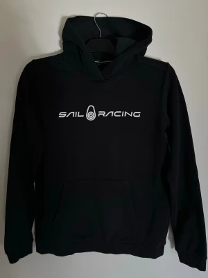 Svart hoodie från Sail Racing - Svart hoodie från Sail Racing med vit logga och text på bröstet samt logga på huvan. Klassisk känguruficka framtill och ribbade muddar vid ärmar och nederkant. Mjuk och bekväm, perfekt för en avslappnad stil. Storlek: 170 cm