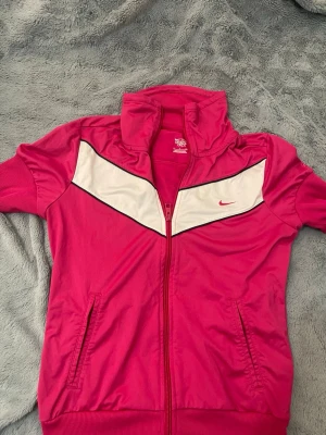 Rosa ziptröja från Nike - Snygg rosa ziptröja från Nike med vit och svart detalj över bröstet. Tröjan har hög krage, ribbade muddar och två fickor framtill. 