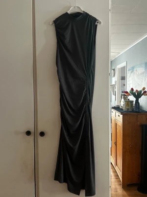 Svart långklänning med drapering - Säljer en svart långklänning med hög hals och ärmlös design. Klänningen har snygg drapering längs sidan som ger en cool och elegant look. Perfekt för dig som vill sticka ut med en stilren och trendig outfit.